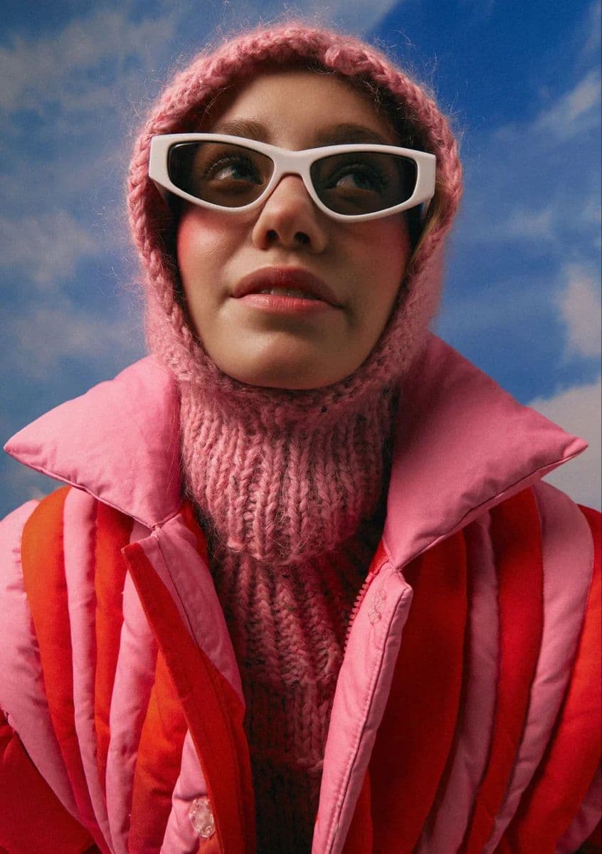 Editorial portrait with pink winterwear and white sunglasses against blue sky